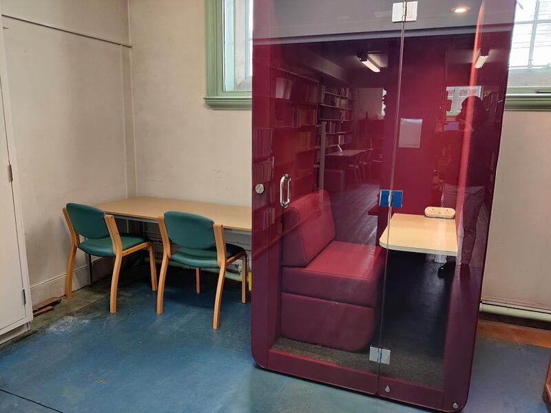 An single occupancy acoustic booth with a small table, power points and a comfy set. The booth is maroon in colour and has a glass door so the occupant is visible.