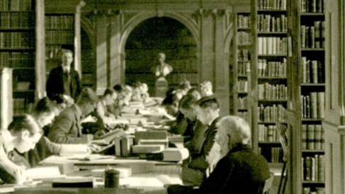 An old black and white photograph of lots of men in traditional Cambridge gowns studying and reading intensively at some old wooden tables which used to run down the centre of the Haddon Library. They are surrounded by oak shelves filled with books.
