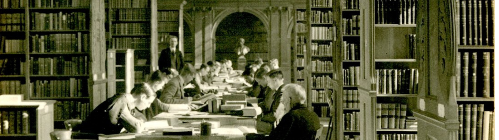 An old black and white photograph of lots of men in traditional Cambridge gowns studying and reading intensively at some old wooden tables which used to run down the centre of the Haddon Library. They are surrounded by oak shelves filled with books.